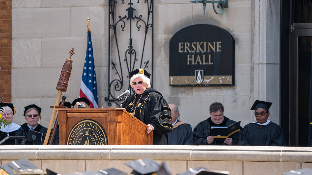 President Connie Gallaher gives opening remarks from a podium on the porch in front of Erskine Hall; members of the ODU faculty and guest speakers sit behind her. 