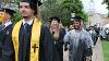 ODU graduates smiling and holding degrees, some in rain ponchos, walk toward the camera. Erskine Hall can be seem framing them in the background.